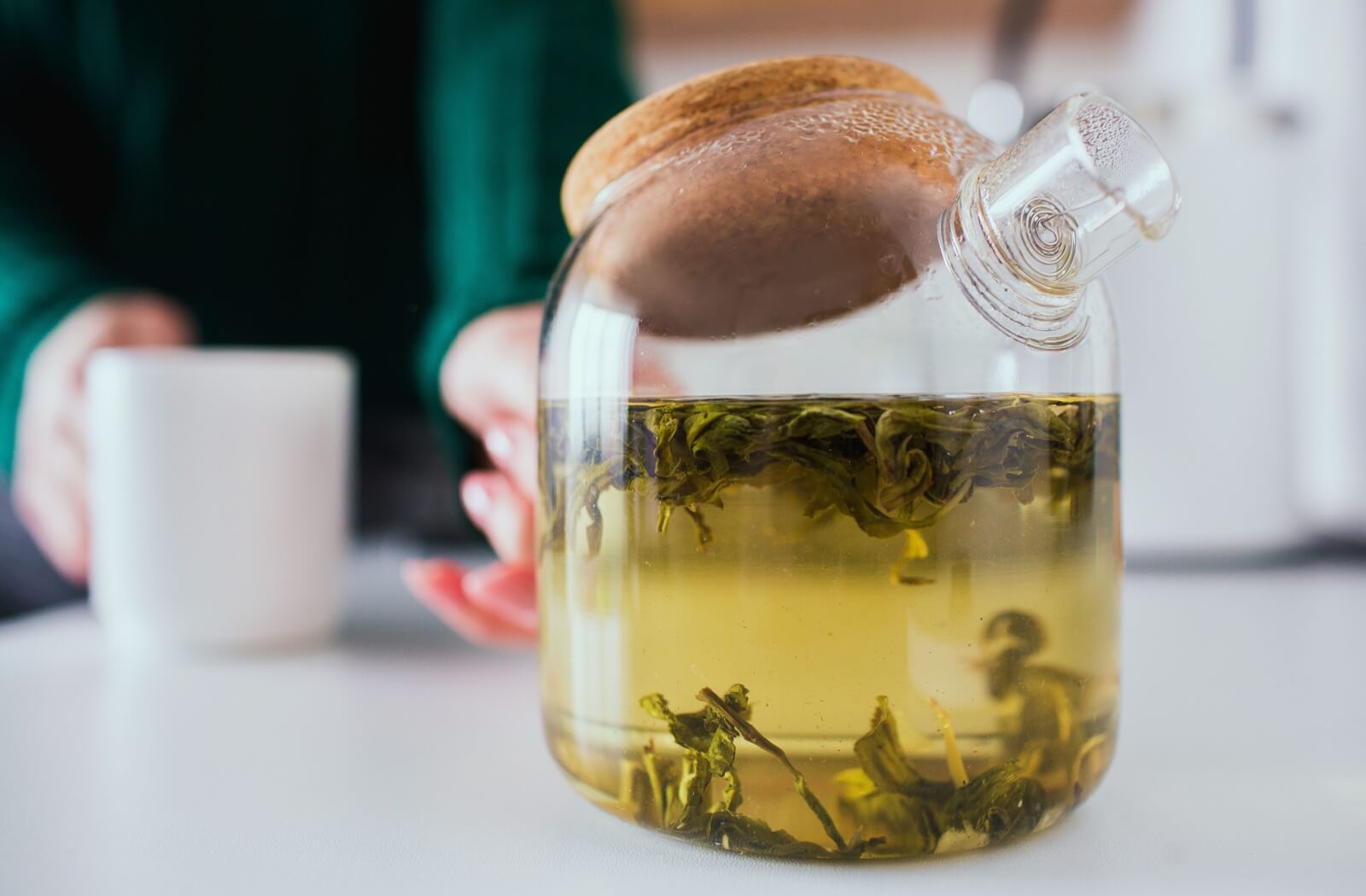 Green tea steeps in a clear pot, someone reaching for it while holding an empty cup.