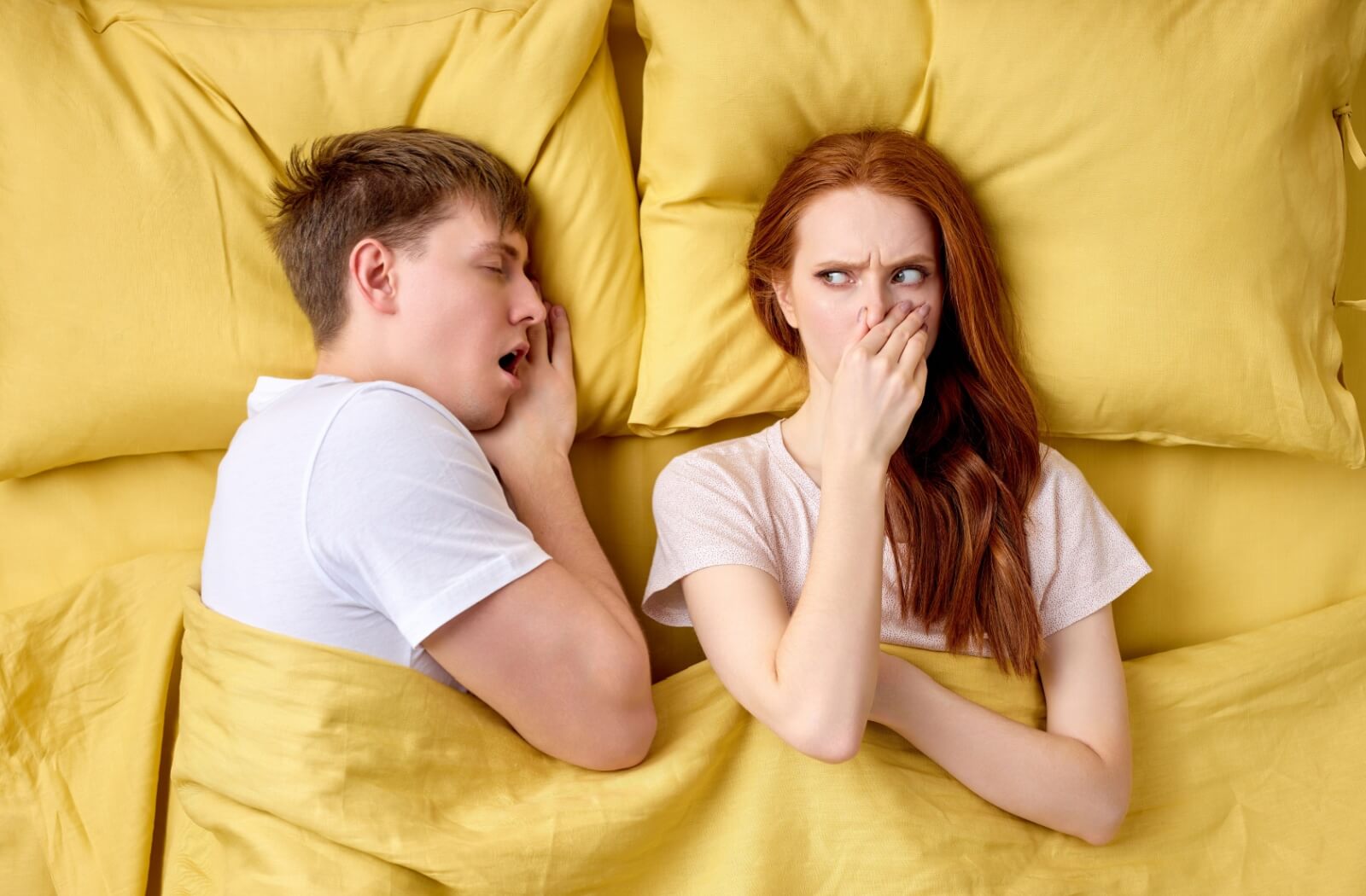 A person lay disgusted in bed next to their partner, who has their mouth open and has bad breath.